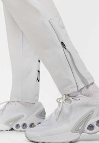 Hvide træningsbukser har sidelukninger og sort branding. De er parret med hvide sneakers, der har grå accenter og synlig luftdæmpning.