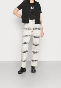 Kvadrovaná černá bezrukávová vesta přes černé tričko, spárovaná s krémovými kalhotami s černými tie-dye pruhy. Černé kotníkové boty.