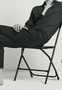 Svart knapp-up skjorta med en lätt glans, mörka jeans med synlig sömnad, sittande på en svart hopfällbar stol mot en vit bakgrund.