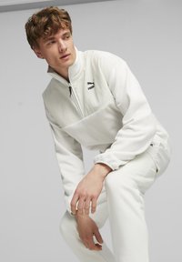 Pullover bianco in pile con colletto alto, chiusura a zip e logo nero. Abbinato a pantaloni bianchi coordinati; texture liscia e vestibilità comoda.