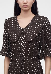 Femme portant une robe noire à pois blancs, avec un grand col à volants et des manches courtes à volants, nouée à la taille.