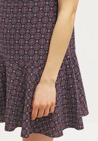 Abito in tessuto blu navy con orlo svasato, caratterizzato da un motivo geometrico rosso e blu. Primo piano di una mano appoggiata sulla gonna.
