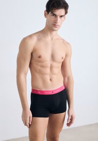 Boxers en coton noir avec une ceinture élastique rose, arborant le nom de la marque "TOMMY HILFIGER" imprimé en blanc. Design simple, texture sans couture.