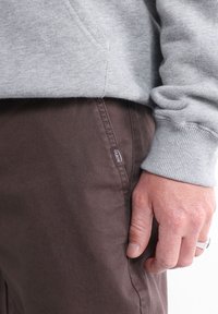 Sweat à capuche en coton gris avec poignets côtelés, associé à un pantalon marron avec une étiquette. Une main visible repose contre la couture latérale du pantalon.