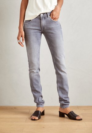 Persoon draagt lichtgrijze slim-fit jeans en zwarte open-toe mules met blokhak, staand op een houten vloer tegen een effen muur.
