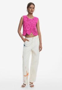 Háčkovaná crop top v žiarivej ružovej s kvetinovými vzormi; biele široké nohavice s farebnými vyšívanými detailmi morského života; biele sandále na remienky.