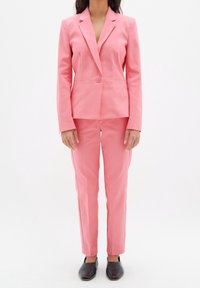 Růžový upravený oblek se sako na jedno knoflíček a klopovými límci, doplněný slim-fit kalhotami. Hladká textura, kombinováno s černými botami.