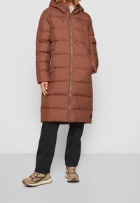 Brun puffjacka med huva, med horisontella kviltade mönster, en framdragkedja och två sidofickor. I kombination med svarta byxor och beiga sneakers.