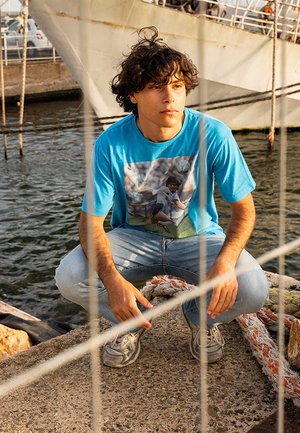 Blauwe t-shirt met een grafische print, lichtblauwe jeans en grijze sneakers. De achtergrond toont water en een schip, met een hek op de voorgrond.