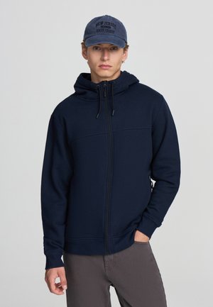 Felpa con zip di colore blu navy, dotata di cappuccio sagomato, cordino e tasche laterali, abbinata a un cappellino blu navy con la scritta "New Jersey East Coast".