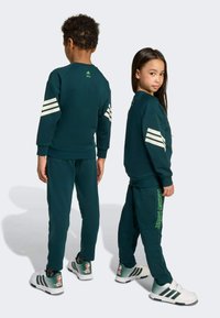 Dark green sweatshirt and pants set with white stripes on sleeves, featuring "Mickey Mouse" print in green. White shoes with colorful accent patterns.