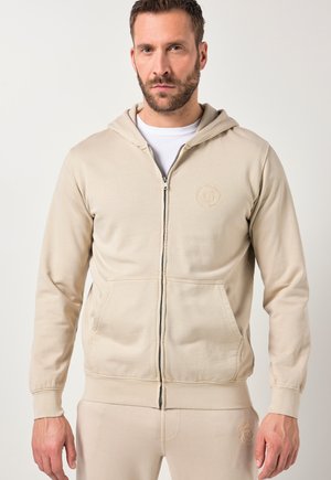 Mand iført beige hættejakke med lynlås og matchende joggingbukser med præget logo, stående foran en ensartet lys baggrund.