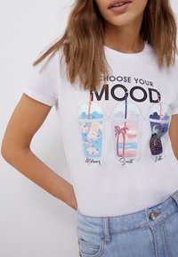 Kvinde iført en hvid t-shirt med teksten "Vælg dit humør" og tre illustrerede kopper mærket Morgen, Solnedgang og Nat med matchende scener.