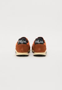 Brune suède og oransje nett sneakers med en svart hælflik som har hvit "VEJA" logo. Kremfarget såle og teksturert svart yttersåle.