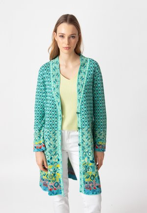 Turkizna kimono jakna s geometrijskim uzorkom, koja ima šarene detalje na manžetama, u kombinaciji s svijetlozelenim topom i bijelim hlačama.