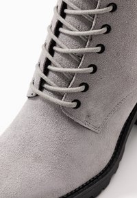 Bota de ante gris claro con puntera redonda, que presenta ojales negros y cordones grises. La suela es texturizada y tiene una ligera elevación.