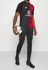Equipación de fútbol que presenta una camiseta negra y roja con rayas horizontales, pantalones negros, logo de Puma y un balón de fútbol en la mano.