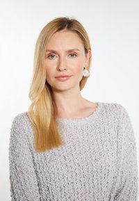 Světle šedý pletený svetr s texturovaným, volným tkaním. Dlouhé rovné blonďaté vlasy, velké kulaté náušnice s růžovým nádechem.