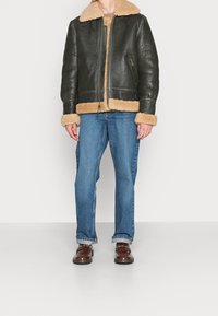 Zwarte leren bomberjack met een tan schapenvacht kraag en manchetten. Gecombineerd met blauwe jeans en bruine schoenen, staande tegen een witte achtergrond.