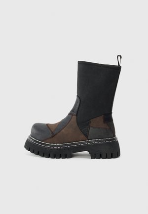 Bota de parcheo en negro y marrón, hasta la mitad de la pantorrilla, con suela gruesa de taco y una asa en la parte trasera, mostrada en perfil lateral sobre un fondo blanco.