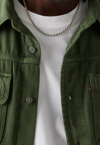 Giacca di cotone verde con bottoni metallici, dotata di una tasca anteriore. Indossata sopra una maglietta bianca, evidenziando trame e colori stratificati.