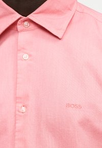 Κοντινό πλάνο ενός ροζ πουκαμίσου με γιακά, λευκά κουμπιά και ένα διακριτικό κεντητό λογότυπο "BOSS" στο ύψος του στήθους.