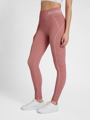 Kvinde iført lyserøde højttaljede sømløse leggings med geometriske mønstre, hvide sneakers og en kort hvid top, står i profil.