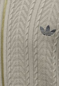 Μπεζ πλεκτό ύφασμα με σχέδιο ψαροκόκαλο, χρυστό φερμουάρ αριστερά και σκοτειλό κεντημένο λογότυπο Adidas Trefoil δεξιά.