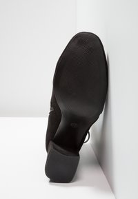 Zapato de ante negro que muestra la suela con goma texturizada para agarre, un tacón afilado y acentos cosidos a lo largo de los lados. Talla visible: 37.