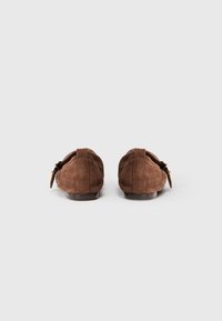 Mocasines de ante marrón con un diseño cosido y detalles de hardware en tono dorado en el lado, presentando un tacón bajo y punta redondeada.