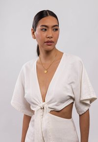 Weißes Leinentop mit kurzen  Ärmeln, geknoteter Vorderseite und tiefem V-Ausschnitt; getragen mit einer goldenen Kettenhalskette mit einem runden Anhänger.