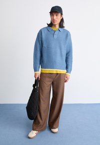 Απαλό μπλε υφασμάτινο πουλόβερ με κίτρινο τελείωμα, καρό καφέ παντελόνι σε φαρδιά γραμμή, άσπρα παπούτσια με κόκκινες λεπτομέρειες και μια μαύρη τσάντα.