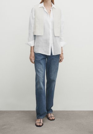 Femme portant une chemise blanche à boutons, un gilet en maille crème posé sur les épaules, un jean bleu et des sandales noires ouvertes, debout sur un sol gris.