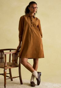 Braunes Leinenkleid mit Knopfleiste, langen Ärmlen und gefältelten Details. Das Model trägt cremfarbene Socken und braune Slipper, neben einem Holzstuhl.