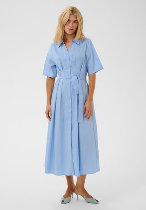 Femme portant une robe chemise ceinturée bleu clair à manches courtes et des chaussures à talons bleu clair, debout sur un fond blanc uni.