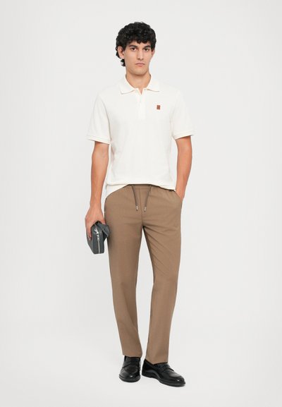 Lys beige polo skjorte med et minimal logo, parret med brune skræddersyede bukser. Modellen holder et mørkegråt tilbehør. Sorte sko fuldender looket.