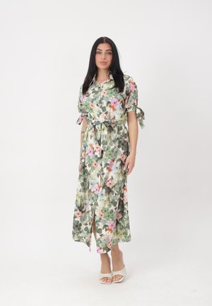 Frau mit langen dunklen Haaren, die ein midilanges Kleid mit Blumenmuster und Gürtel trägt, mit kurzen Puffärmeln und weißen offenen Sandalen, steht vor weißem Hintergrund.