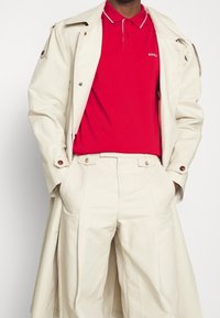 Beige trenchcoat över en röd pikétröja med grå krage, kombinerad med beige veckade byxor. Noterbara knappdekorationer på kappa.