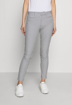 Vrouw draagt slim-fit grijs-witte geruite broek, witte sneakers en een wit mouwloos topje tegen een effen achtergrond.
