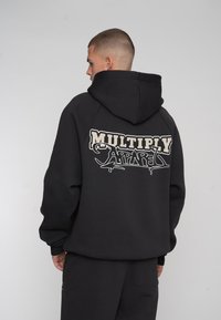 Schwarzer Kapuzenpullover mit oversized Passform, mit weißem Drucktext "MULTIPLY APPAREL" auf der Rückseite. Glatter Stoff, minimalistischem Design.