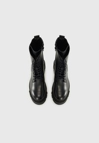 Botas de combate de cuero negro con una suela de goma gruesa, cordones frontales y un zipper lateral para facilitar su uso. Textura suave con un diseño resistente.