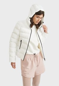 Doudoune blanche avec capuche, design matelassé, fermeture éclair noire et poches latérales, portée sur un sweat-shirt de couleur claire et un short rose.