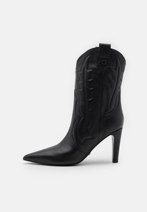 Botte cowboy en cuir noir mi-mollet à talon, avec bout pointu, surpiqûres décoratives et languette de tirage ornée d'un logo "G" en métal.