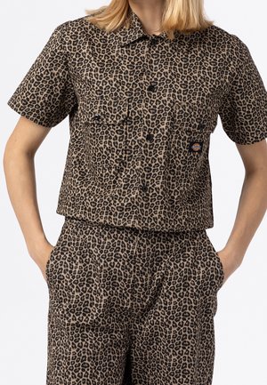 Person trägt kurzärmliges Hemd mit Knöpfen und Hose mit passendem braun-schwarzem Leopardenmuster, Hände in den Hosentaschen.