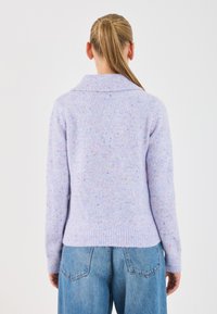 Maglione lilla lavorato a maglia con colletto e polsini a coste, caratterizzato da un motivo maculato in vari colori, indossato con jeans blu a vita alta.