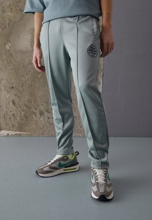 Osoba nosząca jasnoszare spodnie dresowe Nike Athletic Club oraz szare buty sportowe Nike z zielonym logo, stojąca na gładkiej podłodze wewnątrz budynku.