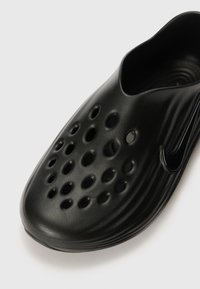 Fekete, formázott, slip-on cipő, több kör alakú szellőzőnyílással, sima textúrával és alacsony profilú kialakítással, az oldalán a Nike logójával.