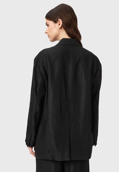 Femme aux cheveux bruns portant un blazer noir ample et un pantalon assorti, de dos sur un fond clair uni.