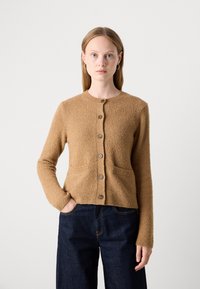 Giovane donna con capelli lisci, che indossa un cardigan beige con bottoni e jeans blu scuro, in piedi con una mano in tasca su uno sfondo bianco.