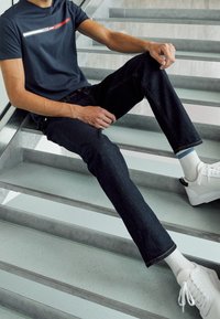 Námořnické bavlněné tričko s horizontálním červeným a bílým vzorem, spárované s tmavými džínami a bílými teniskami se šedými podrážkami.